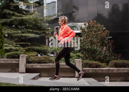 Ein dynamisches Foto mit einer jungen Frau, die in einer Stadt joggt, in einer leuchtend orangefarbenen Jacke und Kopfhörern, zeigt Fitness und Entschlossenheit Stockfoto