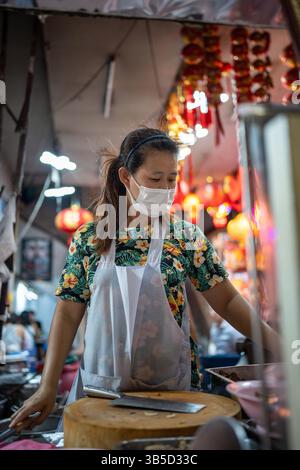 Thailändische Frau mit Gesichtsmaske, die lokale Speisen am Imbissstand in der Yaowarat Road in Chinatown kocht. Stockfoto