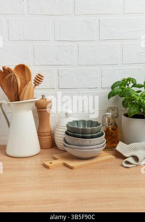 Verschiedene Küchenutensilien, darunter Schüsseln, Holzlöffel, Pfeffermühle, Ölflaschen, und eine Basilikumpflanze, die ordentlich auf einer hölzernen Arbeitsplatte angeordnet sind Stockfoto