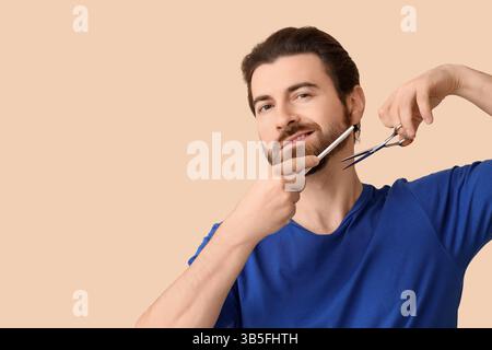 Hübscher junger Mann mit Kamm und Schere, der seinen Bart auf beigefarbenem Hintergrund stylt Stockfoto