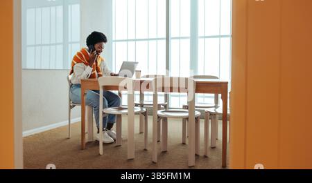 Eine afrikanische Frau saß an einem Holztisch in einem gut beleuchteten Bürobereich, multitasste, indem sie einen Geschäftsanruf tätigte und an ihrem Laptop arbeitete. Die Einstellung ist Stockfoto