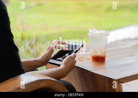 Aufnahme einer Frau, die sitzt und ihr Smartphone benutzt, mit einem Glas Eiskaffee auf dem Tisch nach dem Drink. Das Koffeinkonzept. Kopierbereich. Stockfoto
