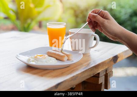 Die Frau isst nach der Gabel und frühstückt auf dem Tisch. Amerikanisches Frühstück mit Spiegeleiern, Würstchen, Kaffee und Saft. Das Konzept eines gesunden Mornins Stockfoto