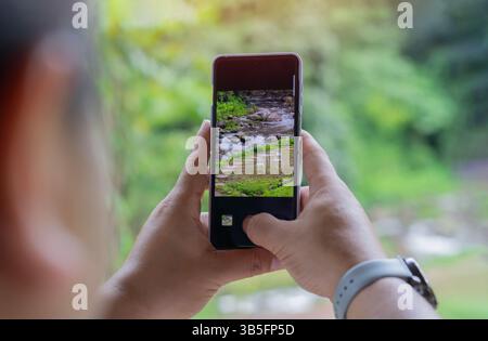 Rückansicht eines nicht erkennbaren männlichen Touristen, der mit seinem Smartphone einen Wasserfall im Wald fotografiert. Selektiver Fokus. Stockfoto