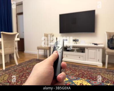 Männliche weiße Hand, die den Fernseher mit schwarzer Plastikfernbedienung in der Wohnung anschaltet Stockfoto