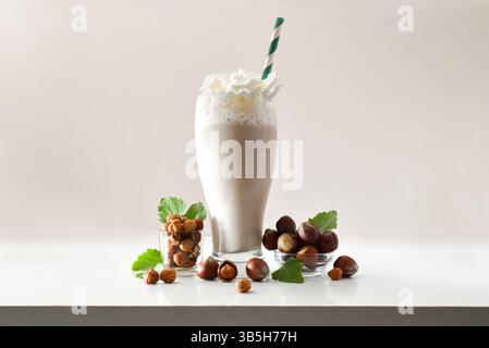 Glas mit Haselnuss-Smoothie mit Creme und grünem Stroh auf weißem Tisch mit Schüsseln voller Haselnüsse und Blätter auf isoliertem hellen Hintergrund. Frontscheibe Stockfoto