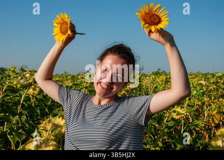 Fröhliche weiße Erwachsene feiert in einem pulsierenden Sonnenblumenfeld unter einem klaren blauen Himmel, hält zwei Sonnenblumen und lächelt hell. Stockfoto