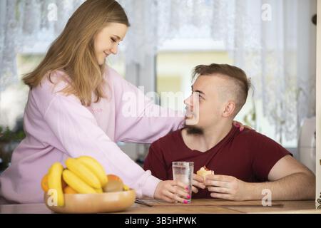 Eine lächelnde blonde Frau mit einem rosa Sweatshirt steht in der Küche. Ein Mann mit Bart sitzt am Tisch und isst ein Sandwich Stockfoto
