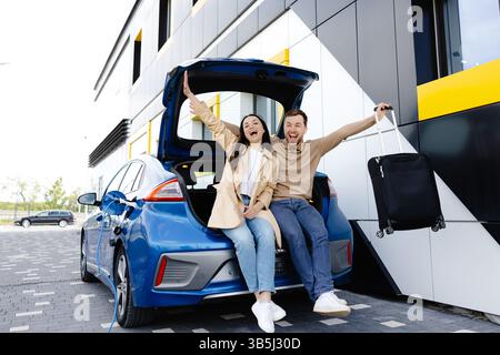 Junge Paare, Mann und Frau, die mit einem Elektroauto unterwegs sind, halten an der Ladestation. Freund und Freundin sitzen im Kofferraum und lächeln. Stockfoto