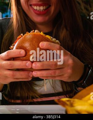 Nahaufnahme einer lächelnden Frau, die einen großen Burger mit verschiedenen Belägen in beiden Händen hält, die eifrig darauf blickt, ihn zu essen. Stockfoto