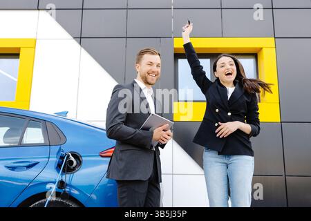 Glückliche Kundin, die vor einem brandneuen Auto steht und die Schlüssel vom Händler im Außenbereich des Händlers abnimmt. Die Frau ist sehr zufrieden mit dem neuen Auto, das sie hält Stockfoto