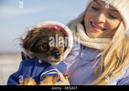 Eine junge blonde Frau lächelt, als sie ihren kleinen Hund ansieht. Sie hält den Hund in ihren Armen. Die Frau und der Hund sind draußen, während es eiskalt ist Stockfoto