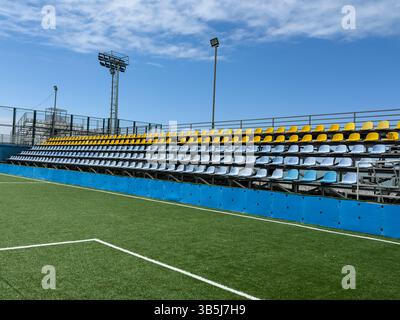 Leere farbenfrohe Stadionsitze und Kunstrasen-Fußballfeld unter klarem blauem Himmel. Städtische Sportinfrastruktur, keine Leute, Outdoor-Umgebung. Stockfoto