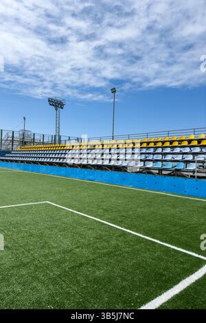Vertikale Ansicht der leeren Stadionstände mit farbigen Sitzen und Kunstrasen-Fußballfeld unter blauem Himmel. Sportstätte im Freien, keine Leute. Stockfoto
