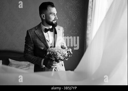 Ein stilvoller Bräutigam in einem gemusterten Anzug hält einen Brautstrauß und blickt nachdenklich aus dem Fenster. Das Zimmer hat elegante Tapeten. Monochromatisches Bild. Stockfoto