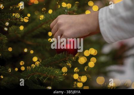 Eine Hand platziert ein glänzendes rotes Ornament auf einem wunderschön verzierten Weihnachtsbaum mit leuchtenden Bokeh-Lichtern Stockfoto