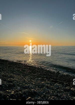Kieselstrand bei Sonnenuntergang mit ruhigem Meer und Sonnenspiegelung auf dem Wasser. Sanfte Farben, natürliches Licht, friedliche Küstenlandschaft, keine Leute, Abendstimmung. Stockfoto