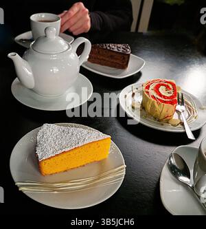 Oben ein Bild von einem Tisch mit einer weißen Teekanne, zwei Tassen Tee und drei verschiedenen Scheiben Kuchen: Ein gepuderter gelber Kuchen, ein dekorierter gerollter Kuchen, ein Stockfoto