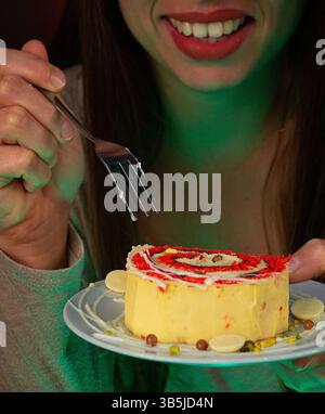 Nahaufnahme einer lächelnden Frau, die einen Teller mit einem dekorierten Stück Kuchen hält und dabei ist, einen Biss mit einer Gabel zu nehmen. Stockfoto
