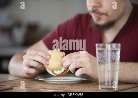 Ein Mann sitzt an einem Tisch. Vor ihm steht ein Teller und ein Glas Wasser. Er hält ein Gemüse-Sandwich in der Hand. Unscharfer Hintergrund Stockfoto