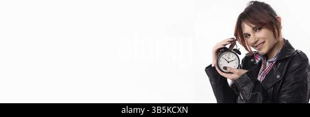 Eine fröhliche junge Frau in einer Lederjacke hält einen Wecker und symbolisiert Zeitmanagement und modernes Leben Stockfoto