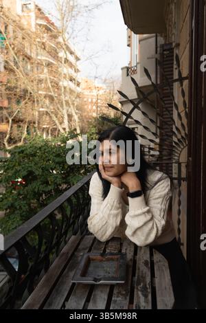 Eine Frau sitzt auf einem Balkon und blickt auf die Stadt. Sie trägt einen weißen Pullover und hat eine Zigarette in der Hand Stockfoto