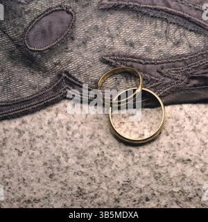 Zwei elegante goldene Ringe liegen auf einer strukturierten Spitze- und Marmoroberfläche, die ihren Glanz zum Ausdruck bringen. Dieses Arrangement weckt Liebesgefühle. Stockfoto