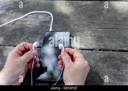 Hände, die weiße Ohrhörer an ein Smartphone auf einem Holztisch anschließen, um Musik oder Podcasts zu hören Stockfoto