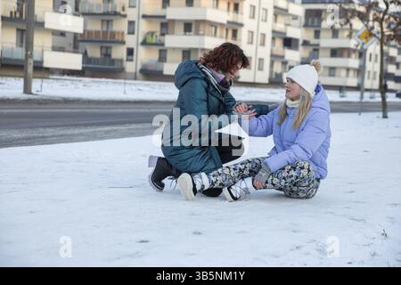 Ein Mädchen versucht, ihrem Freund beim Aufstehen zu helfen. Der Freund ist umgefallen und sitzt im Schnee. Sie hat sich im Herbst gequetscht und hält sie fest Stockfoto