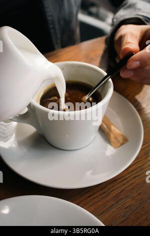 Mädchen, das Milch in eine Kaffeetasse gießt. Hände, die Milch mit einem Löffel in Kaffee rühren. Latte im Café. Espresso mit Milch. Heißgetränke-Konzept. Entspannen Sie sich bei einem Kaffee. Stockfoto