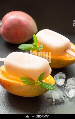 Mangosorbet-Eis auf halb geschnittenen Mangobrüchten hinter Mango in der Nähe von Eiswürfeln auf schwarzem Hintergrund. Mangobruchtsorbet-Eis-Eis. Vertikal. Stockfoto