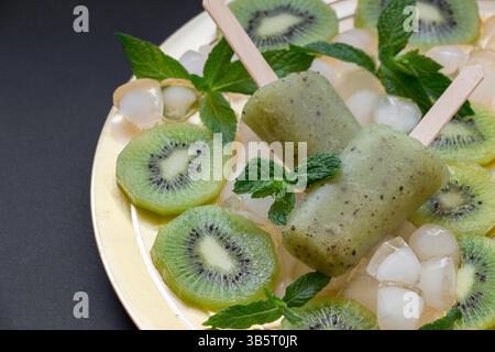 Kiwi-Sorbet-Eis-Eis auf goldener Platte mit Eiswürfeln gehackten Kiwi-Scheiben und Minzblättern auf schwarzem Hintergrund. Kiwi-Sorbet-Eis popsic Stockfoto