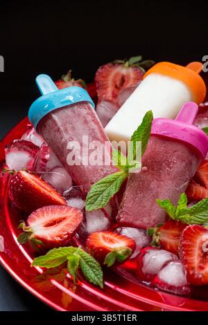 Erdbeereis Eisbecher auf rotem Teller mit Eiswürfeln gehackte Erdbeeren und Minzblätter auf schwarzem Hintergrund. Erdbeereis Sonne Stockfoto