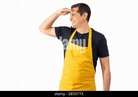 Verkäufer trägt gelbe Schürze über isoliertem weißem Hintergrund sehr glücklich und lächelnd und blickte weit weg mit Hand über Kopf. Suchkonzept. Stockfoto