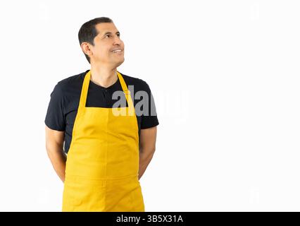 Lateinischer Mann mit gelber Schürze auf weißem Hintergrund schaut auf den Kopierraum, Lieferdienstmitarbeiter, Kellner in gelber Schürze Stockfoto