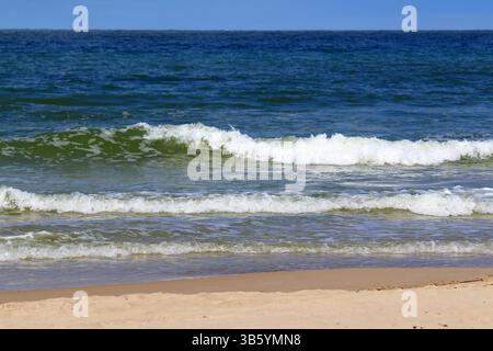 Wellen, die auf einen Sandstrand der Ostsee unter klarem blauem Himmel Rollen, weite Sicht auf die pulsierende Küste und das offene Meer bei natürlichem Tageslicht. Stockfoto