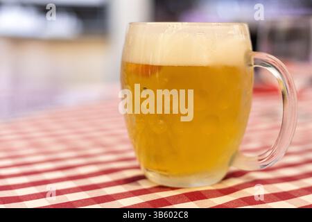 Ein mit goldenem Bier gefüllter klarer Glasbecher liegt auf einer rot-weiß karierten Tischdecke. Um ihn herum bietet eine lebhafte Atmosphäre einen festlichen Abend Stockfoto