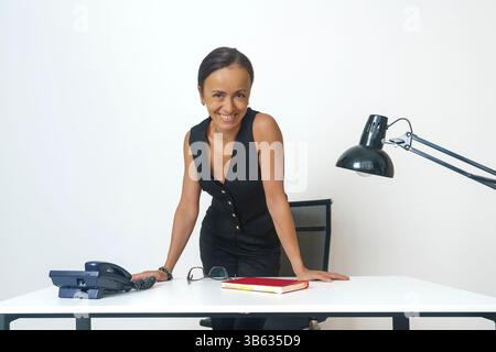 Eine lächelnde Geschäftsfrau in einem schwarzen Anzug steht am Tisch und lehnt sich mit den Händen darauf Stockfoto