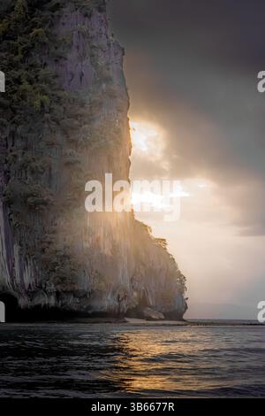 Dieses atemberaubende Bild fängt eine dramatische Meereslandschaft bei Sonnenuntergang ein. Die hoch aufragenden, zerklüfteten Klippen erheben sich aus dem ruhigen, reflektierenden Wasser und werfen lange Schatten Stockfoto
