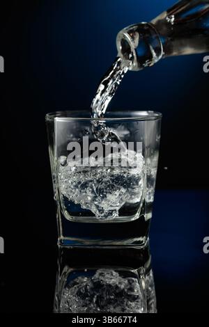 Kristallklarer Wodka oder Gin wird über Eis in ein Glas gegossen. Wodka mit Eis auf schwarzem reflektierendem Hintergrund. Stockfoto