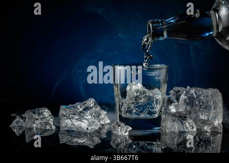 Kristallklarer Wodka oder Gin wird über Eis in ein Glas gegossen. Wodka und Eisstücke auf schwarzem reflektierendem Hintergrund. Kopierbereich. Stockfoto