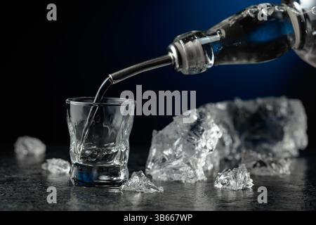 Kristallklarer Wodka oder Gin wird in ein Glas gegossen. Transparentes alkoholisches Getränk mit Eisstücken auf schwarzem Hintergrund. Stockfoto