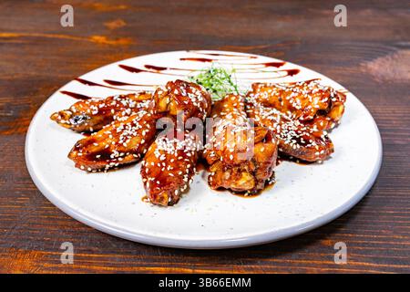 Ein Teller hält köstliche glasierte Hühnerflügel, die mit Sesamsamen bestreut sind, auf einem rustikalen Holztisch. Das Gericht wird kunstvoll mit einem Spritzer Sau präsentiert Stockfoto
