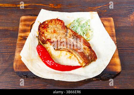 Gebratene Schweinerippchen werden auf einem Holzbrett zusammen mit einer frischen Gemüsegarnitur und einer roten Chili-Paprika präsentiert, was ein schmackhaftes Gericht für lun darstellt Stockfoto