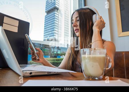 Junge Frau, die in einem Café arbeitet, Laptop und Smartphone benutzt, Kopfhörer trägt und ein Getränk genießt Stockfoto