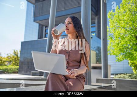 Eine junge Geschäftsfrau trinkt Kaffee und arbeitet an ihrem Laptop vor einem modernen Bürogebäude und genießt eine produktive Pause Stockfoto