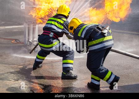 Feuerwehrleute, die gelbe Helme und Schutzanzüge tragen, arbeiten zusammen, um einen Großbrand mithilfe eines leistungsstarken Wasserschlauchs zu kontrollieren und zu löschen Stockfoto