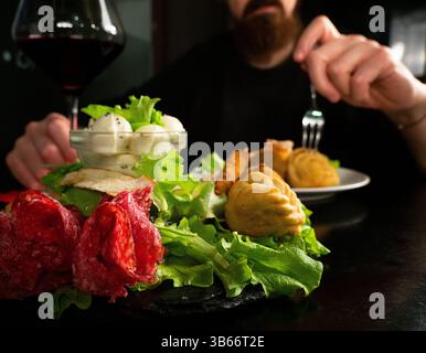 Nahaufnahme der Hände eines Mannes, der eine Gabel über einem Teller mit Vorspeisen hält, mit einem Glas Rotwein in der Nähe. Eine Schüssel Mozzarella und Salat ist in der Stirn Stockfoto