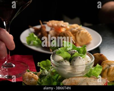 Nahaufnahme einer Hand mit einem Glas Rotwein neben einem Teller mit verschiedenen Gourmet-Vorspeisen und einer Glasschale mit Mozzarella und Salat. Stockfoto