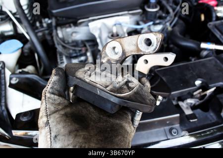 Austausch der Motorhalterung, Nahaufnahme einer mechanischen Hand mit Handschuhen, die eine abgenutzte Motorhalterung des Fahrzeugs hält. Fahrzeugwartung. Stockfoto